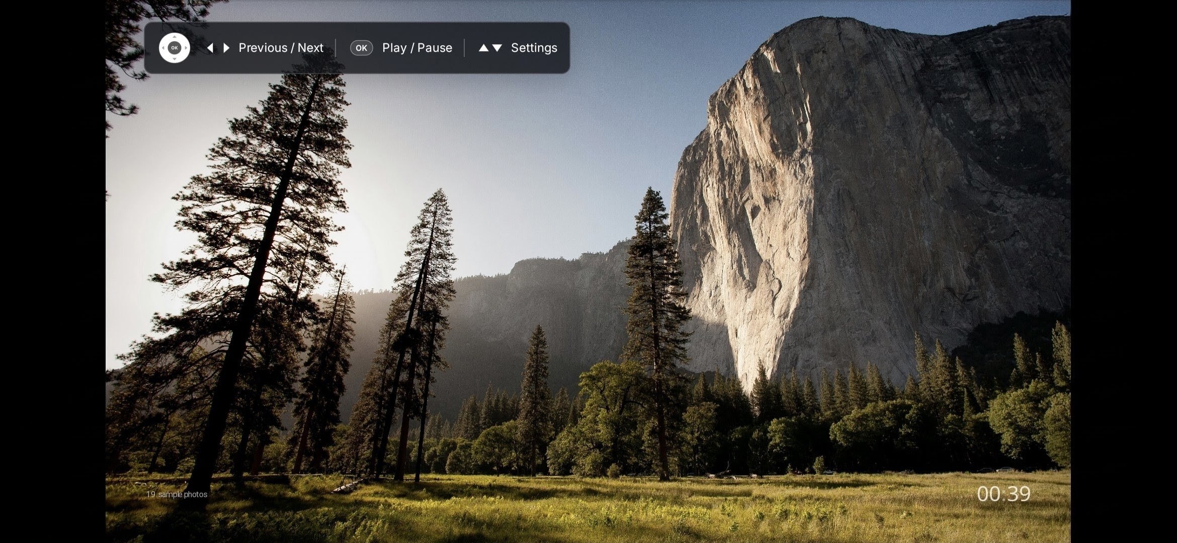 PictaCast displaying a mountain landscape on a TV, with a compact hint showing Previous / Next, OK for Play / Pause, and Up / Down for Settings